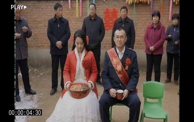 A still from a video (timestamp 00:00:04:30) showing a Chinese wedding ceremony with a bride in a white dress holding a red basin and a groom wearing a '新郎' (groom) sash, surrounded by family members in a rural setting.