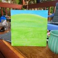 A small green notebook or notepad rests on a wooden table next to a teal mug, sitting on what appears to be an outdoor patio.