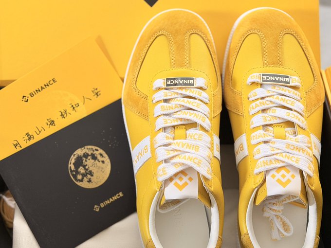 A pair of yellow sneakers with white laces featuring the 'BINANCE' logo, displayed alongside a yellow Binance-branded box that includes Chinese characters reading '问海山海·秋中入学'.