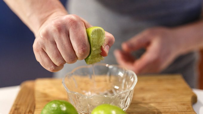 A close-up of hands squeezing lime juice into a glass bowl on a wooden cutting board, with whole limes visible.