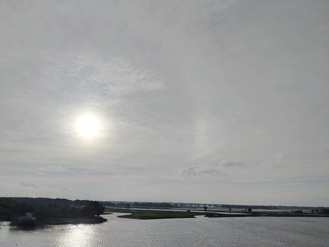 A scenic outdoor photograph showing a bright sun in a cloudy sky over a wide river with green islands and a distant bridge.