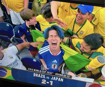 A FIFA World Cup 2026 Round of 32 match between Brazil and Japan (2-1), showing an enthusiastic Japanese fan with blue hair celebrating alongside Brazilian fans in yellow.