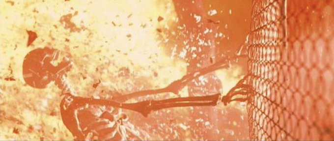 The image shows a dramatic scene of a skeleton's arm reaching toward a chain-link fence amid a fiery explosion, symbolizing desperate circumstances. It visually represents the post's portrayal of Coinbase users pleading with CEO Brian Armstrong for a lifeline, framing their request for unregged tokens as a do-or-die plea akin to disarming a catastrophic threat.