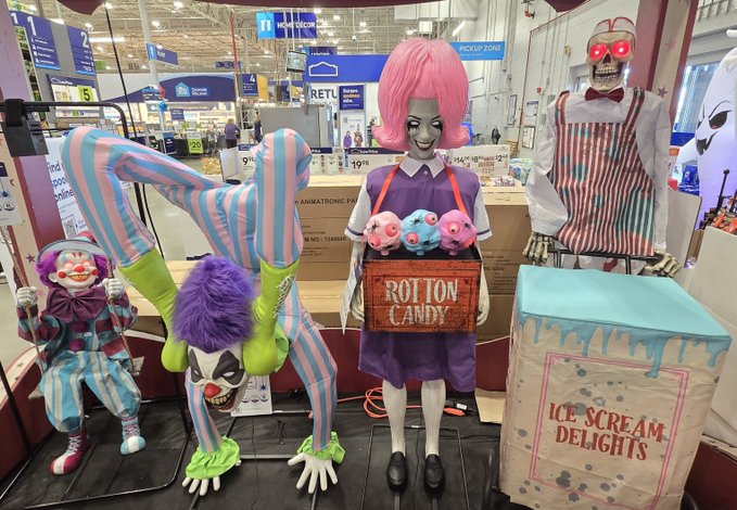 A display of spooky clown and zombie Halloween decorations at a store, including hanging clown figures, a mannequin with a pink wig holding a "COTTON CANDY" crate, and a box labeled "ICE SCREAM DELIGHTS."