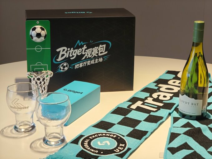 A soccer-themed "Bitget" viewing kit is displayed, featuring a branded box, a miniature net with a soccer ball, a teal "5UBU" case, two etched glasses, a checkered scarf with "Firdaus" and "UEX" text, and a bottle of Cloudy Bay wine. The arrangement of merchandise and drinks complements the post about receiving a World Cup viewing package from Bitget, suggesting a celebratory setup for watching matches.