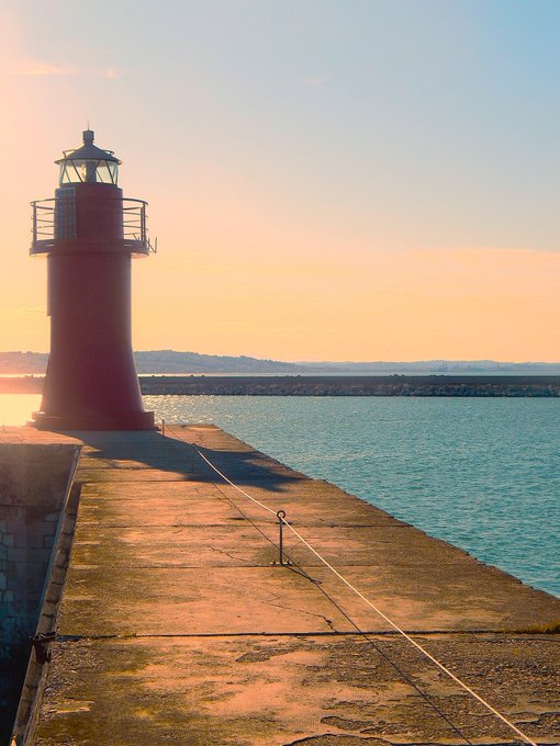 A scenic photograph of a red lighthouse on a concrete pier at sunset, overlooking calm blue water with a distant shoreline.