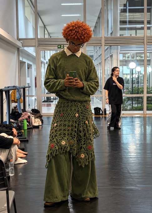 A person with bright orange curly hair, wearing an olive green knit sweater, crocheted skirt, and wide-leg pants, stands in an indoor space looking down at their phone.