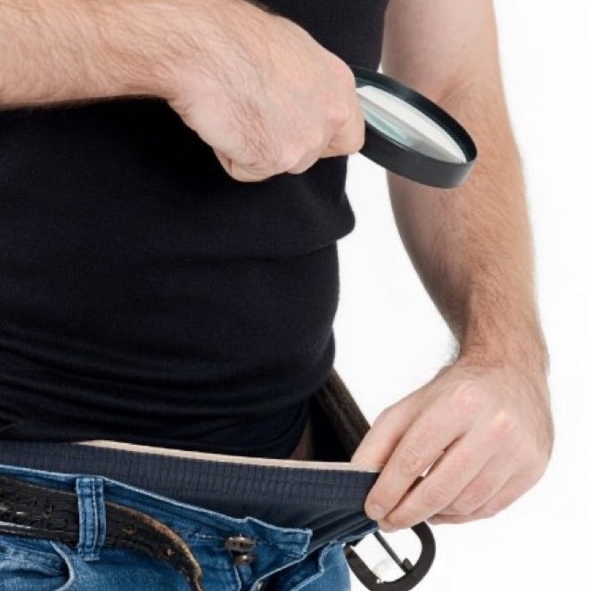A man examining his stomach with a magnifying glass while pulling up his shirt, revealing his belly over his jeans.
