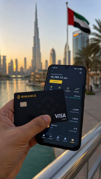 A hand holds a black Binance Visa debit card in front of a smartphone displaying a wallet balance of 10,789.12 AED, with the Dubai skyline (including the Burj Khalifa) and a UAE flag in the background.