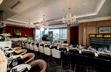 An elegant, dimly-lit restaurant interior features crystal chandeliers, white-clothed dining tables, plush chairs, and a long banquette with gold decorative panels.