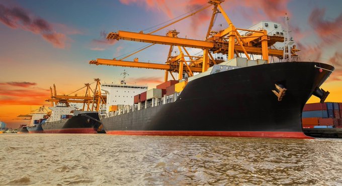 A large black cargo ship loaded with shipping containers is docked at a port with orange cranes, set against a dramatic orange sunset sky.