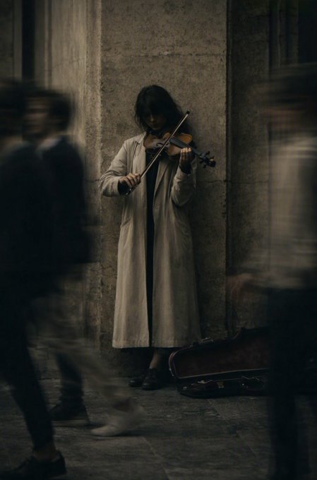 A woman in a long coat stands playing a violin against a weathered stone wall as blurred figures of pedestrians walk past her on a city street.