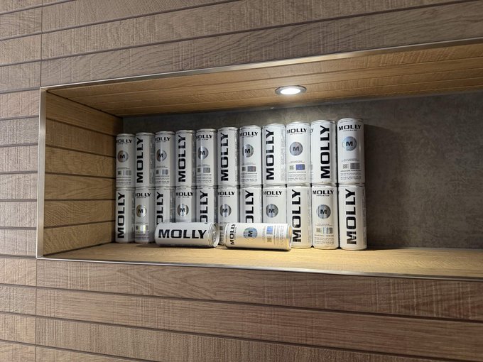 A shelf displays stacks of white "MOLLY" branded cans (resembling energy drink packaging) arranged neatly in a recessed wooden display with accent lighting. The image appears to be a humorous metaphor for the post's theme, as "Molly" is slang for MDMA, playfully referencing the author's frustration and "losing confidence" in Base following controversial actions by Jesse Pollak and Brian Armstrong.