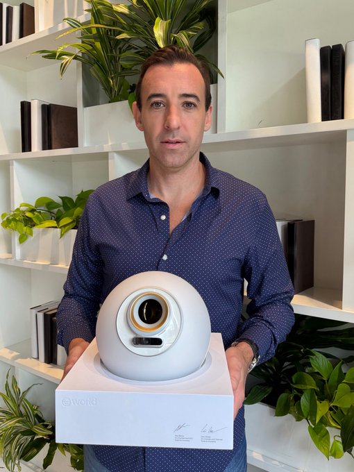 A man wearing a blue shirt stands next to a white box containing an orb, which he holds up for the camera. The image also includes text that reads "finally received my Orb! scanning your eyeball to prove you're human onchain. the identity layer for every human on earth is being built in real time." This suggests that the man might be involved in a project related to facial recognition or identity verification, possibly using the orb as part of the process.