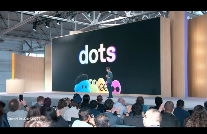 A photo from OpenAI DevDay [2025] showing a presenter on stage in front of a large screen displaying the word 'dots' and colorful blob characters, with an audience watching.
