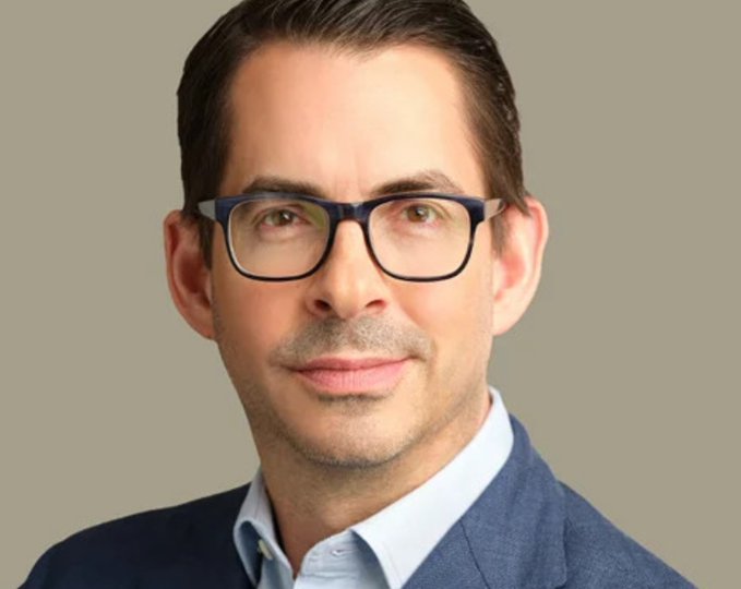 A professional headshot of a smiling man with dark hair wearing glasses and a blue suit jacket over a light blue shirt.