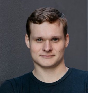 A headshot photograph of a young man with short light brown hair wearing a dark blue t-shirt against a gray background, smiling slightly at the camera.