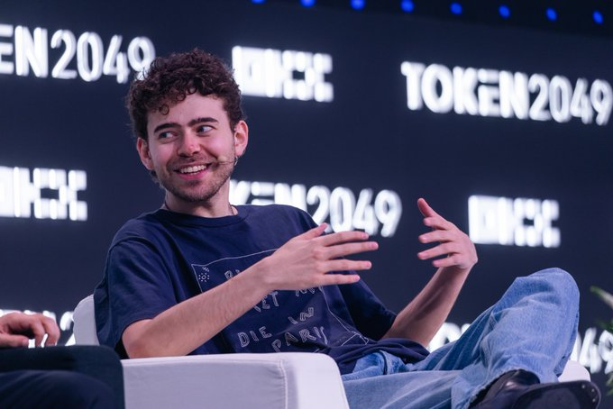A young man with curly brown hair sits on a panel at the "TOKEN2049" conference, smiling and gesturing with his hands.
