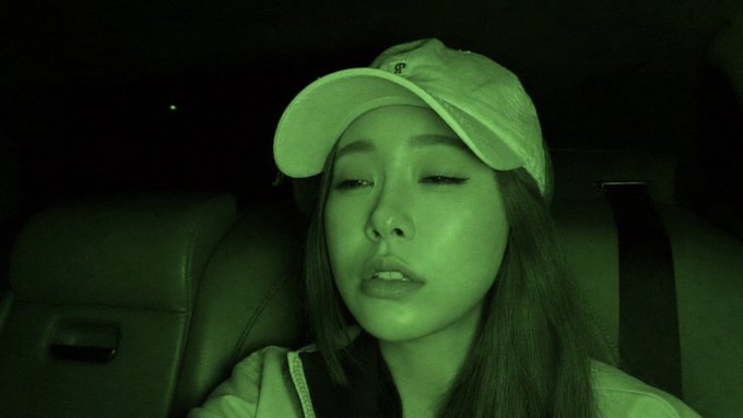A young woman wearing a white cap poses for a selfie in a car at night, with the image having a green night-vision filter effect.