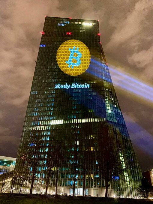 A skyscraper displays a light projection at night featuring a Bitcoin logo and the text 'study Bitcoin'.