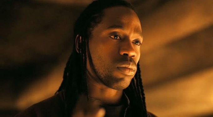 A dramatic close-up of a man with dreadlocks looking thoughtfully off-camera in warm, golden lighting.