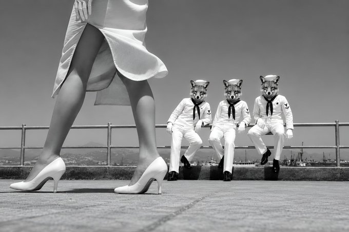The image features three people wearing sailor uniforms, with one person standing behind them and two others sitting down. The person standing behind them has their legs crossed over a railing, while the other two individuals are seated at the edge of the scene. The background includes a fence or barrier that separates the viewer from the sailors.