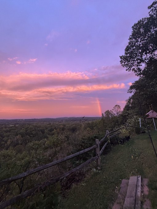 A scenic sunset view from a wooden fence-lined overlook, featuring pink and purple clouds, a faint rainbow arcing over distant hills, and rolling green forest below.
