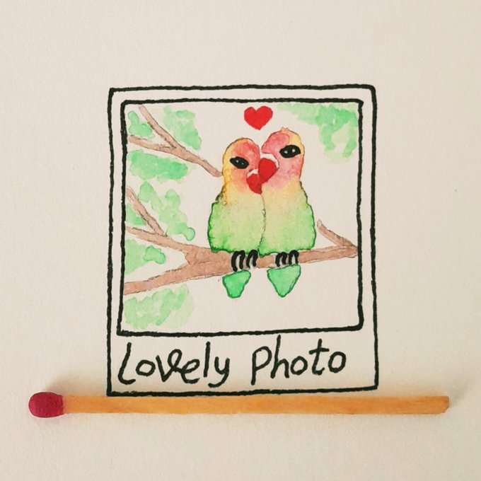 A hand-drawn watercolor illustration of two colorful lovebirds on a branch with a red heart above them, framed in a 'Lovely Photo' border, with a wooden matchstick placed below for scale.