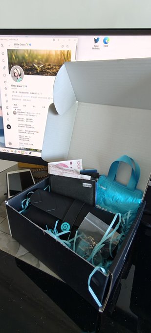 The image features a black box with a blue ribbon tied around its neck, sitting on top of a table. The box contains various items such as a wallet, a handbag, and some other small objects. In the background, there is a computer monitor displaying an image of a bird. The post includes a caption that reads "一大早被 Bitget 的惊喜礼盒治愈了！ 质感直接拉满，大清早开箱心情都变好了。新的一天继续充满动力", which seems to be a reference to the post about Bitget.