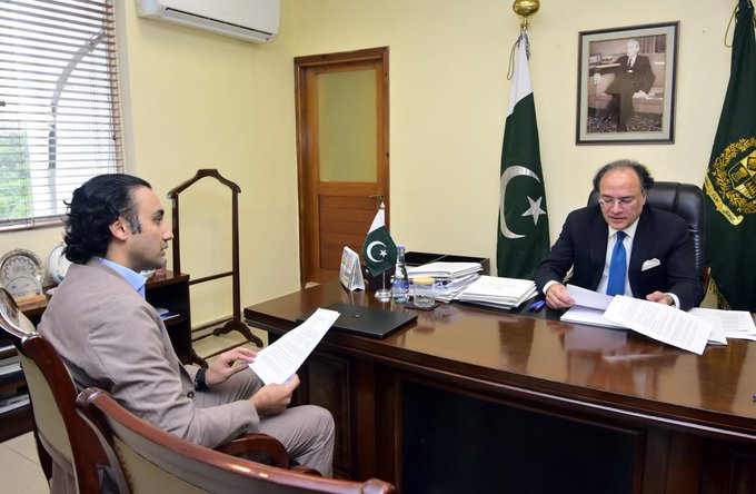 Finance Minister Senator Muhammad Aurangzeb reviews documents during a meeting at his office with PVARA Chairman Mr. Bilal Bin Saqib, who briefed him on insights from his recent Hong Kong visit. The Pakistani flag and a framed portrait of the national founder are visible in the background of the ministerial office.