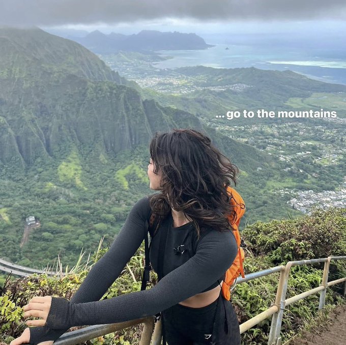 A woman with dark hair in athletic wear and an orange backpack sitting at a scenic mountain overlook with the text '... go to the mountains'.