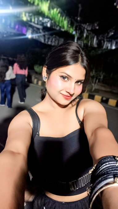 A young woman with dark hair pulled back takes a nighttime selfie wearing a black sleeveless top with metallic buckle details, smiling at the camera with other people and trees visible in the background.