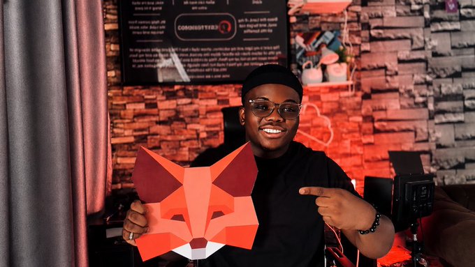 The image features a man wearing glasses holding up an orange paper fox mask, which has the words "Web3" written on it. The man appears to be smiling for the camera while posing with the mask. The background of the photo shows a brick wall, and there is also a couch in the scene.