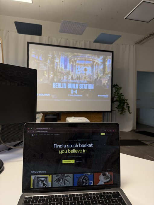 A workspace showing a laptop displaying the 'Finary' website ('Find a stock basket you believe in') in the foreground, with a projector screen in the background showing 'BERLIN BUILD STATION'.