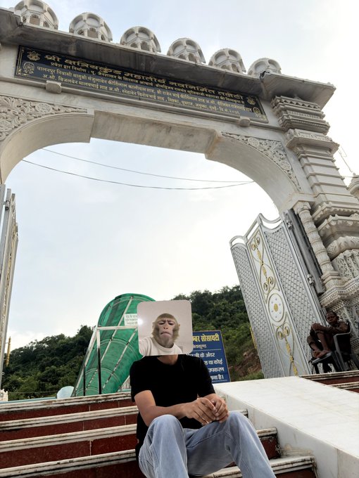 A person sits on stone steps beneath a large, ornate archway featuring Hindi script, while a small inset image of a monkey in a white shirt floats above their head.