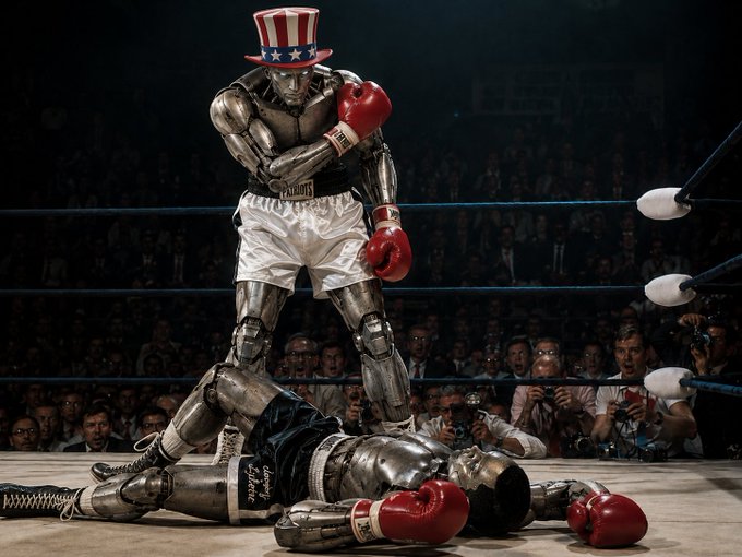 A humanoid robot wearing a patriotic Uncle Sam top hat and red boxing gloves stands victorious over a defeated opponent in a boxing ring, with a crowd watching in the background.