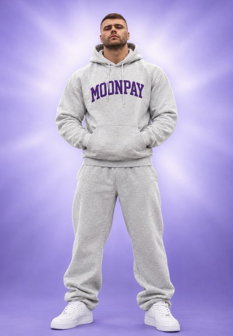 A man wearing a gray 'MOONPAY' hoodie and matching sweatpants stands with hands in pockets against a purple cloudy background, wearing white sneakers.