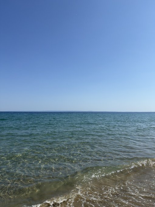 A calm ocean scene with clear blue water, gentle waves lapping at a sandy shore, and a vast cloudless blue sky above the horizon.