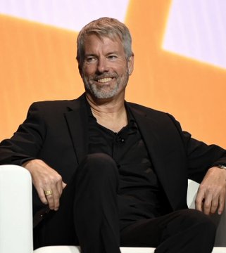 A smiling, gray-bearded man in a black suit sits relaxed in a white chair against an orange backdrop, likely at a corporate or investor event. The image appears to show Michael Saylor, the executive chairman of Strategy (formerly MicroStrategy), whose company just raised $2.01 billion by selling 18.26 million $MSTR shares to bolster its Bitcoin treasury strategy with $1.59 billion in cash.