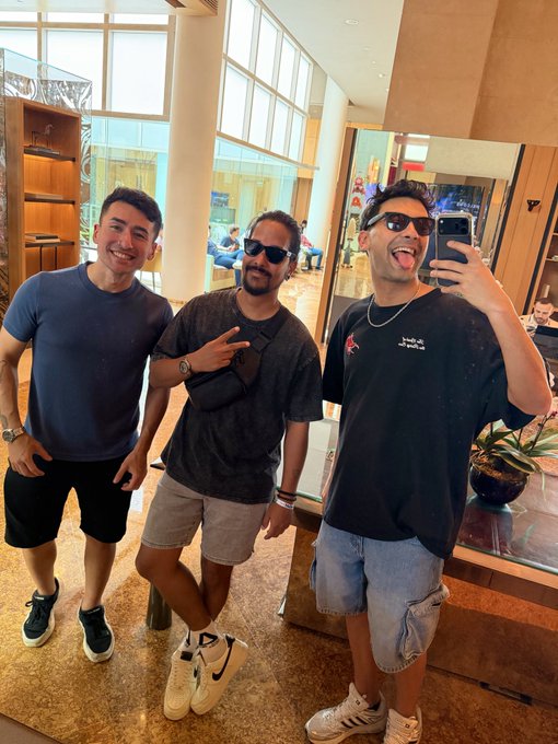 Three young men posing for a selfie in a lobby or hotel setting, with the person on the right wearing a shirt that has Arabic text reading 'يلا نمشي' (Let's go).