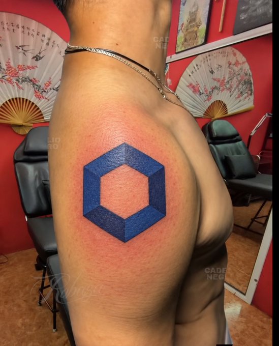A close-up view of a person's upper arm featuring a fresh tattoo of a blue geometric hexagonal cube design, set inside a tattoo parlor decorated with fans and artwork.