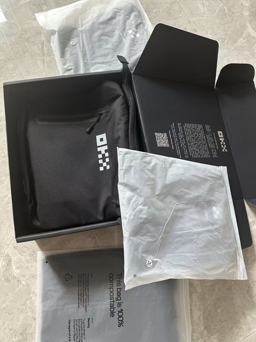 An unboxing photo showing an OKX-branded black box containing a black OKX zippered pouch/bag, with additional white packaging materials including a compostable bag labeled 'This bag is 100% compostable.'