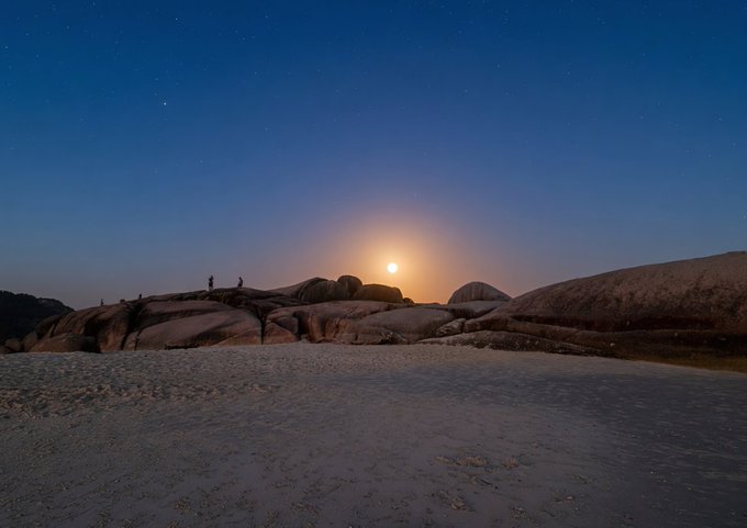 A nighttime landscape photograph showing the moon setting over large rock formations with small silhouettes of people, viewed across a sandy beach under a starry sky.