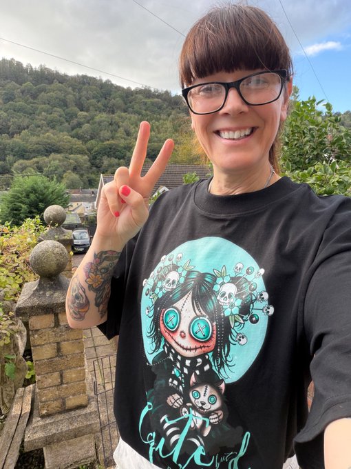 A smiling woman with short brown hair and glasses wearing a black t-shirt featuring a sugar skull design flashes a peace sign, with a wooded hillside visible behind her.