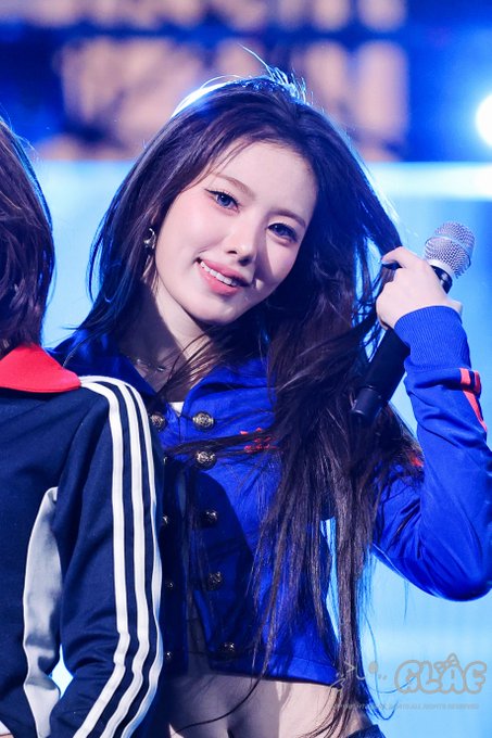 A young woman with long dark hair wearing a blue jacket holds a microphone while smiling on stage.