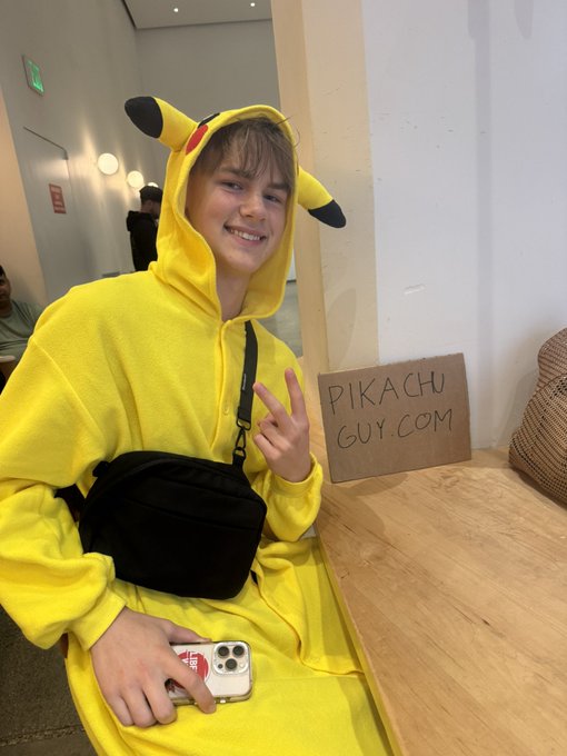 A photograph of a smiling young man wearing a yellow Pikachu onesie, holding a phone and giving a peace sign, seated next to a handwritten cardboard sign that reads 'PIKACHU GUY.COM'.