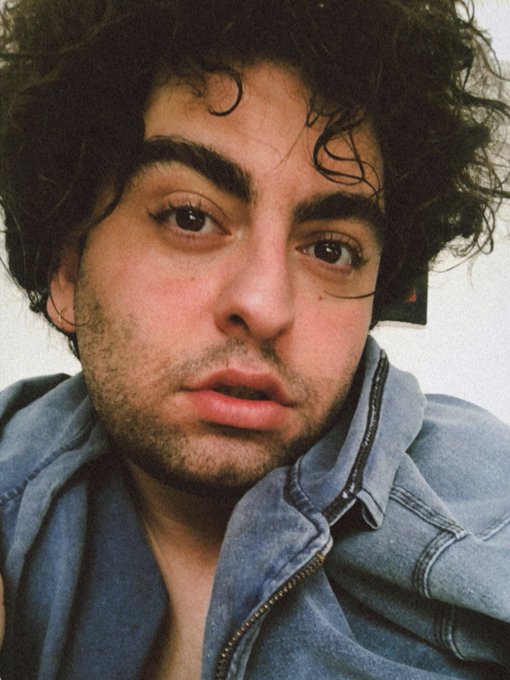 A close-up selfie of a young man with curly dark hair, prominent eyebrows, and facial stubble wearing a blue denim jacket.