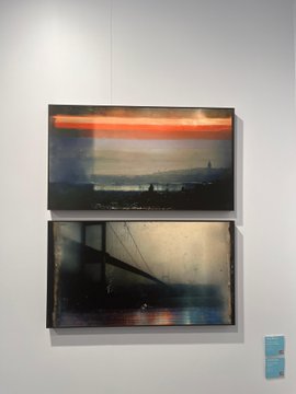 Two framed artworks hang on a white gallery wall, depicting dark, atmospheric landscapes with a prominent orange/red band of light across the top of the upper piece and a bridge silhouette in the lower piece.