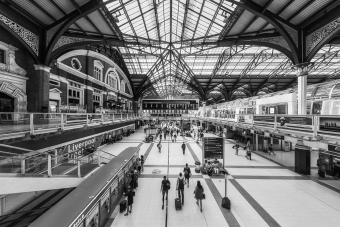 Black and white photograph of Liverpool Street Station showing travelers with luggage beneath an ornate arched glass roof.