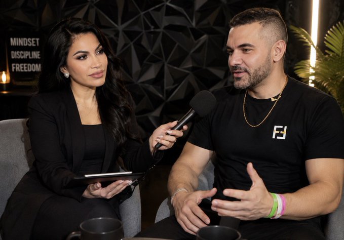 A woman with long dark hair, wearing a black blazer, holds a microphone up to a bearded man in a black t-shirt who is speaking with animated hand gestures during what appears to be a podcast recording. The setting features a dark geometric wall with the visible text "MINDSET DISCIPLINE FREEDOM," and the scene aligns with the post's caption about creating a premier crypto podcast aimed at cleaning up the industry.